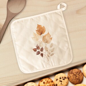 May include: A beige cotton pot holder with a design of brown and beige leaves. The pot holder has a loop for hanging.