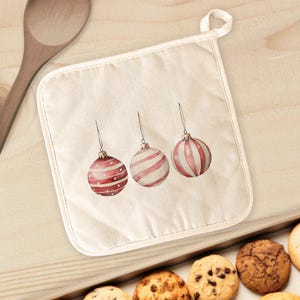 May include: Off-white pot holder featuring three watercolor Christmas ornaments in red and white. The ornaments have different designs, including stripes and polka dots. A loop is attached for hanging. Cookies are visible in the background.