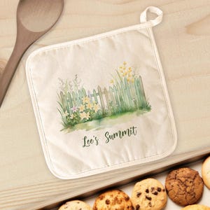 May include: A white cotton pot holder with a watercolor illustration of a picket fence, flowers, and greenery. The text "Lee's Summit" is printed below the illustration.