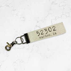 May include: A beige canvas keychain with the text "52302 marion, ia" printed in black. The keychain has a bronze-colored clasp and key ring.