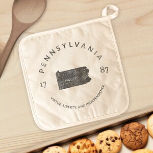 May include: A white cotton pot holder with a black print of the state of Pennsylvania, the words "Pennsylvania 1787 Virtue, Liberty, and Independence" and a black outline of the state.
