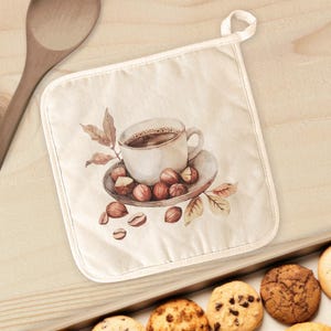May include: A cream-colored pot holder with a watercolor design of a coffee cup, hazelnuts, and leaves. The pot holder has a white trim and a loop for hanging. Cookies are visible in the bottom right corner.