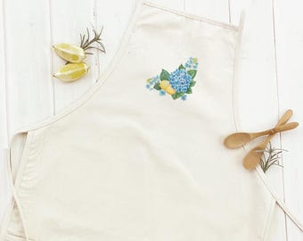 May include: Off-white apron with a floral design featuring blue hydrangeas and yellow lemons. The apron has a front pocket and long ties. Two wooden spoons and lemon halves are placed on a white wooden surface.