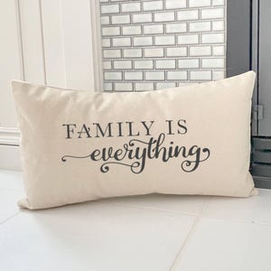May include: A rectangular beige pillow with the phrase "FAMILY IS everything" printed in black script. The pillow is made of a soft fabric and is placed on a white tiled surface.