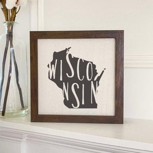 May include: A framed canvas print with a dark brown frame. The canvas features a black silhouette of the state of Wisconsin with the word "WISCONSIN" written in white letters inside the state outline.