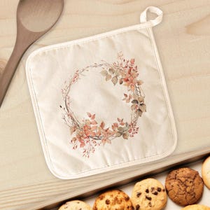 May include: A square, off-white pot holder with a floral wreath design in shades of brown, orange, and green. The pot holder has a loop for hanging and is placed near a wooden spoon and a tray of cookies.