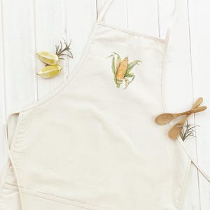 May include: Off-white apron with a watercolor illustration of an ear of corn. The apron has a pocket and neck strap. Two wooden spoons and lemon slices are placed near the apron. The apron is made of a durable fabric.