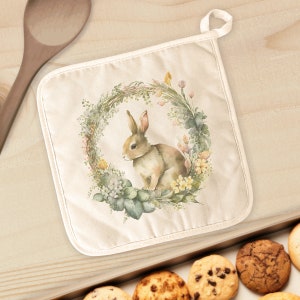 May include: A white cotton pot holder with a floral wreath design featuring a brown rabbit sitting inside the wreath. The wreath is made of green leaves, pink and yellow flowers, and brown twigs.