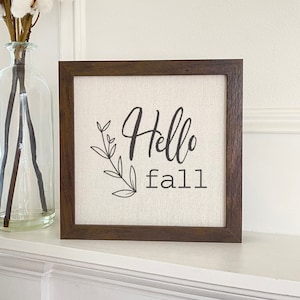 May include: Square wooden framed canvas art with the words "Hello fall" in black script and a leaf design. The canvas has a neutral linen texture. The frame is a dark brown color. The art is displayed on a white surface.
