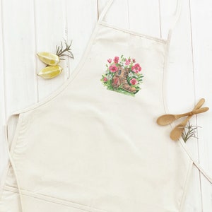 May include: A white cotton apron with a small, colorful design of a house in a boot surrounded by pink flowers.