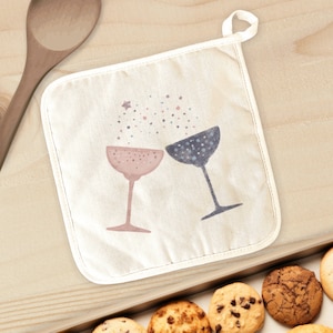 May include: A white cotton pot holder with a design of two champagne glasses clinking together, with pink and blue accents and a scattering of gold stars.