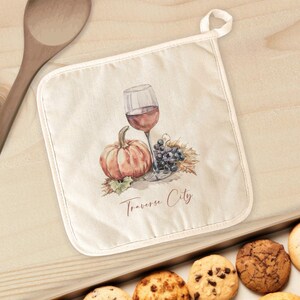 May include: Off-white pot holder with a watercolor design of a wine glass, pumpkin, and grapes. The text "Traverse City" is printed below the image. The pot holder has a white border and a loop for hanging.