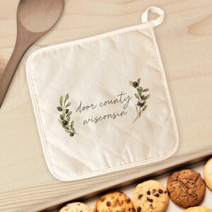 May include: Off-white pot holder with the text "Door County Wisconsin" in a script font, framed by green olive branch illustrations. The pot holder has a white trim and a loop for hanging. Cookies are visible in the foreground.
