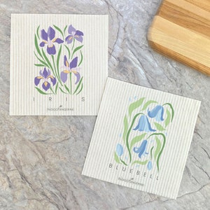 May include: Two kitchen cloths with floral designs. The first cloth features purple irises with yellow centers and green leaves. The second cloth features bluebells with green leaves. Both cloths have the text "INDIGOTANGERINE" printed on them.