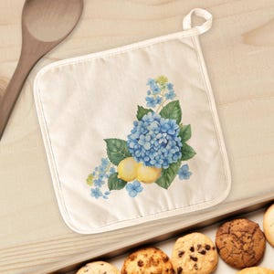 May include: A square, off-white pot holder with a loop for hanging. It features a watercolor design of blue hydrangeas, green leaves, and yellow lemons. The pot holder is on a light wooden surface.