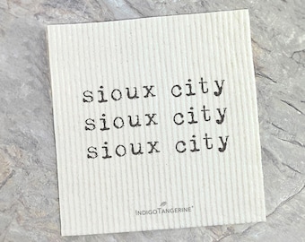 May include: A square, off-white absorbent cloth with the words "sioux city" printed three times in black, typewriter-style font. The brand name "INDIGOTANGERINE" is printed in the bottom right corner.