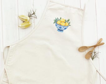 May include: Off-white apron with a blue and white bowl design filled with yellow lemons and green leaves. Two wooden spoons and lemon halves are next to the apron. The apron has a pocket and neck and waist ties.