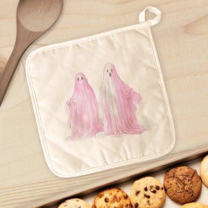May include: Off-white pot holder with a loop, featuring two pink ghost illustrations. The ghosts have open mouths and flowing fabric. The pot holder is on a light wooden surface, with a wooden spoon and cookies visible.