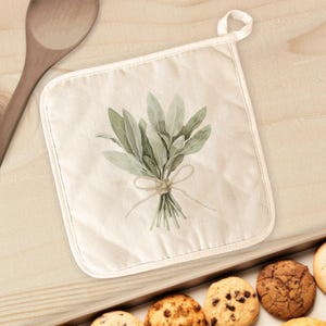 May include: Off-white square pot holder with a watercolor illustration of sage tied with twine. The pot holder has a loop for hanging and is surrounded by a wooden spoon and a tray of cookies.