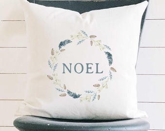 May include: White decorative pillow with the word "NOEL" in blue, surrounded by a wreath of green and blue leaves and brown pinecones. The pillow is on a black chair against a white background.