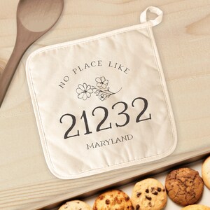 May include: A beige cotton pot holder with black text that reads "No place like 21232 Maryland" and a small floral design.