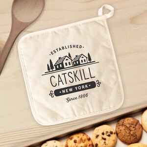 May include: A white cotton pot holder with a black logo that says "Established. Catskill. New York. Since 1806". The logo features a silhouette of two houses with trees.