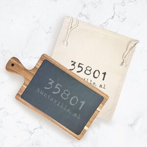 May include: A wooden serving board with a dark gray slate surface, featuring the engraved text "35801 Huntsville, al." A beige drawstring bag with the same text is behind the board. The board has a handle.