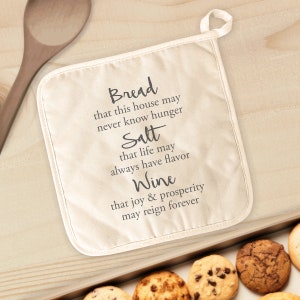 May include: A white cotton pot holder with black text that reads "Bread that this house may never know hunger, Salt that life may always have flavor, Wine that joy & prosperity may reign forever."