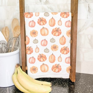 May include: A white kitchen towel with a pattern of orange and brown pumpkins. The towel is hanging on a wooden rack.