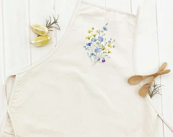 May include: A white cotton apron with a floral design featuring blue and yellow wildflowers. The apron has adjustable straps and a large pocket.