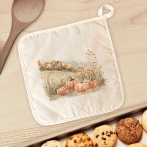 May include: A square, off-white pot holder with a loop for hanging. The center features a watercolor painting of pumpkins in a field with autumn foliage. The pot holder is bordered with a cream-colored trim.