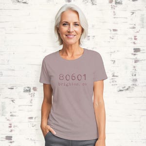 May include: A mauve colored t-shirt with a white crew neck. The shirt has a pink graphic print of the text "80601 brighton, co"