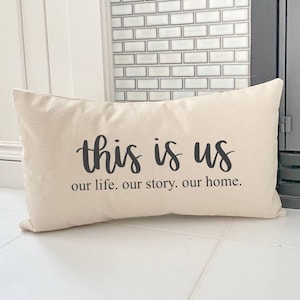 May include: A rectangular beige pillow with the phrase "this is us" in black script, followed by "our life. our story. our home." in a smaller font. The pillow is on a white tiled surface.