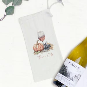 May include: White wine bottle bag with a watercolor illustration of a wine glass, pumpkin, and grapes. The text "Traverse City" is printed below the illustration. A bottle of Chardonnay is partially visible.