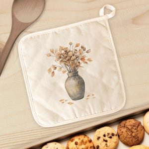 May include: Off-white square pot holder with a watercolor illustration of a gray vase filled with dried flowers. The pot holder has a white trim and a loop for hanging. The background is a light wood surface.