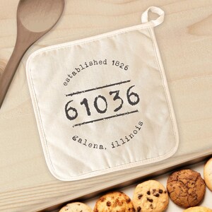 May include: Off-white pot holder with the text "established 1826 61036 galena, illinois" in black. The pot holder has a loop for hanging and is next to a wooden spoon and a tray of cookies.