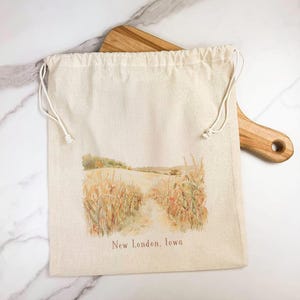 May include: A beige drawstring bag with a watercolor illustration of a rural landscape featuring a path through a field of wheat. The text "New London, Iowa" is printed below the image. The bag is made of a natural fabric.