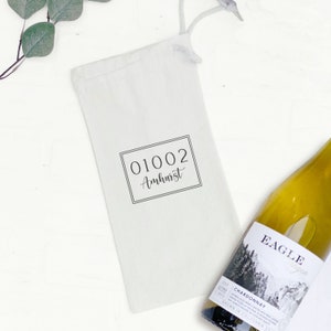 May include: White cotton wine bag with a black drawstring. The bag has a black rectangular label with the text "01002 Amherst" printed on it. A bottle of white wine is next to the bag.