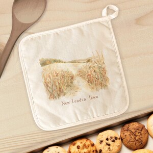 May include: Off-white pot holder with a watercolor image of a field and the text "New London, Iowa". The pot holder has a loop for hanging and is surrounded by a white border. A wooden spoon is visible in the background.