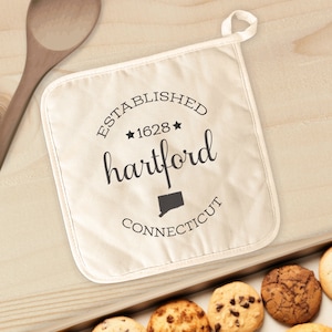 May include: A beige pot holder with the text "ESTABLISHED 1628 HARTFORD CONNECTICUT" in black. The pot holder has a loop for hanging and is next to a wooden spoon and a tray of cookies.