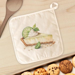 May include: Off-white square pot holder with a watercolor illustration of a slice of key lime pie. The pie has a graham cracker crust, lime filling, whipped cream, and a lime slice. A small loop is at the top for hanging.