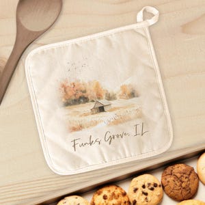 May include: Off-white pot holder with a watercolor landscape of a barn and field, with the text "Funks Grove, IL". The pot holder has a white trim and a loop for hanging. Cookies are in a tray below.