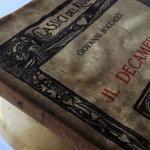 Può includere: Un libro vintage intitolato "Il Decameron" di Giovanni Boccaccio. La copertina del libro presenta un design ornato con un cuore e un'illustrazione incorniciata di una figura con una spada.