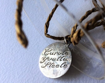 May include: A silver charm with the name "Carole Yvette Steele" engraved on it. The charm is attached to a brown cord and is hanging from a clear glass object.