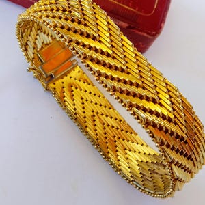 Puede incluir: Una pulsera ancha de color dorado con un diseño tejido. La pulsera presenta una serie de eslabones rectangulares dispuestos en un patrón de chevrón. El cierre es visible y la pulsera está sobre una superficie blanca con una caja roja en el fondo.