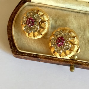 May include: A pair of gold tone flower-shaped earrings with pink and clear rhinestones. The earrings are in a small, brown velvet box.