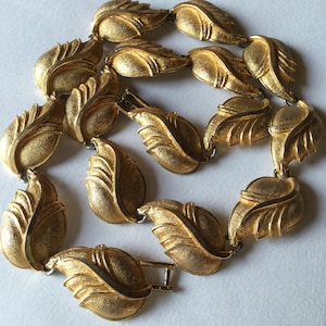 Op de afbeelding: Een gouden ketting met een bladontwerp. Elk blad is getextureerd en heeft een licht verhoogd midden. De ketting is gemaakt van metaal en heeft een sluiting.