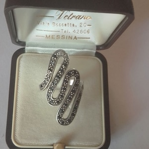 Può includere: Un anello d'argento con un designo di pietra nera a forma di onda, in una scatola di anello di velluto bianco con il testo "Vetrano, Via Boccetta, 20 - Tel. 42806 MESSINA" sul coperchio.