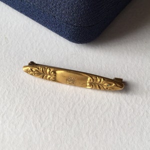 Könnte beinhalten: Eine goldfarbene Bar-Brosche mit einem verzierten Design und einem kleinen gravierten Symbol in der Mitte.