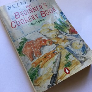 May include: A vintage cookbook titled "Betty Falk, The Beginner's Cookery Book, New Edition". The cover features a watercolor illustration of a wooden cutting board with a knife, carrots, onions, and other vegetables.